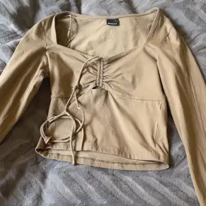 Snygg beige långärmad topp från Gina med knytning framtill och v-ringad design. Perfekt för en stilren look.