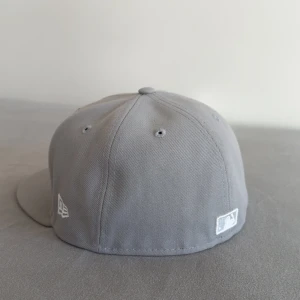 Grå keps från New Era - Snygg grå keps från New Era med broderad vit LA-logga på framsidan. Kepsen har en klassisk 59FIFTY design med en platt skärm och är tillverkad i bomull. Perfekt för en stilren look. Relativt ny - Köptes 2024. Endast använd ett fåtal gånger. Mycket bra skick. Kvitto tillgänglig. 7 1/4 - 57.7cm 
