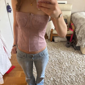 Ljuslila korsett med dragkedja - Säljer en stilren ljuslila korsett med dragkedja framtill och snörning baktill. Den är axelbandslös och har en figurnära passform som framhäver midjan. Perfekt för att ge en elegant touch till din outfit.