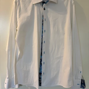 Vit skjorta med blå mönstrade detaljer från Lindbergh - Säljer en vit skjorta från Lindbergh med unika blå och vita mönstrade detaljer på insidan av krage, knappslå och manschetter. Skjortan har lång ärm och klassisk krage, perfekt för dig som vill sticka ut lite extra.