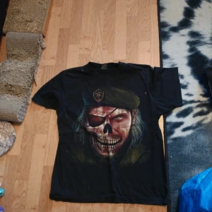 Svart t-shirt med dödskalletryck från Rock Eagle - Cool svart t-shirt från Rock Eagle med stort tryck av en dödskalle med ögonlapp och basker på framsidan. T-shirten är i 100% bomull och har normal passform med korta ärmar. Perfekt för dig som gillar unika och tuffa motiv.