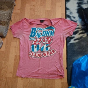 Rosa t-shirt med tryck från Shine - Rosa t-shirt från Shine med stort färgglatt tryck på framsidan: 'The Big DNM Year 62 Jeans Wear' i blått, rött och vitt. T-shirten har korta ärmar och rund hals. Perfekt för en avslappnad stil.