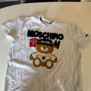 Vit t-shirt med Moschino-tryck - Säljer en vit t-shirt från Moschino med ett stort tryck av en nallebjörn och texten 'Moschino' i svart och rött. T-shirten har korta ärmar och en rund halsringning. Perfekt för en avslappnad stil!