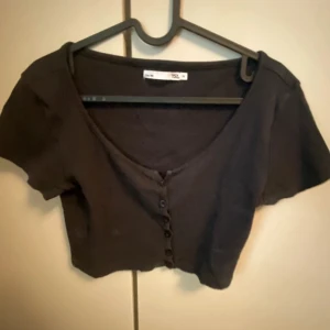 Svart kortärmad topp från 157 - Säljer en svart kortärmad topp från 157 med v-ringning och knappar framtill. Perfekt för en stilren look.