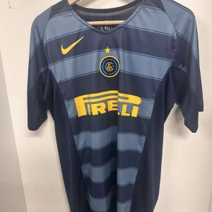 Inter Milan fotbollströja med Adriano 10 - Snygg Inter Milan fotbollströja från Nike med Adriano och nummer 10 på ryggen. Tröjan är randig i mörkblå och ljusblå nyanser med gula detaljer och Pirelli-logga på framsidan. Perfekt för fans av laget och spelaren!