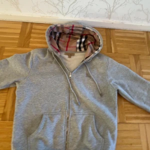 Burberry Grå  - Säljer en snygg grå hoodie från Burberry med dragkedja och ett klassiskt rutigt foder i huvan. Perfekt för en stilren och bekväm look. Den har långa ärmar och en praktisk ficka framtill. Passar perfekt för kyligare dagar.