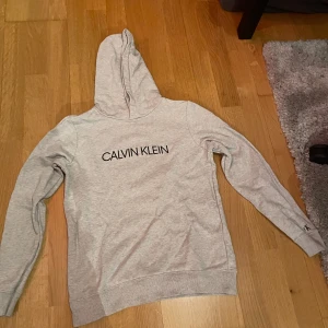 Grå hoodie från Calvin Klein - Säljer en stilren grå hoodie från Calvin Klein med svart logotyp på framsidan. Tröjan har långa ärmar och en praktisk huva. Perfekt för en avslappnad look. Nästan aldrig använd 