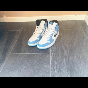 Jordan 1 uneversity blue - Snygga Nike Air Jordans i en fräsch blå och vit färgkombination. Skorna har en hög design med klassisk snörning och den ikoniska Nike-loggan på sidan. Perfekta för att ge din stil ett sportigt lyft.
