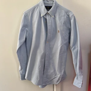 Blå skjorta från Ralph Lauren - Säljer en stilren blå skjorta från Ralph Lauren med broderad logga på bröstet. Skjortan har en klassisk krage med knappar och är långärmad. Perfekt för en snygg och avslappnad look. Mindre storlek, passar främre S