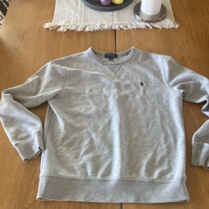 Grå sweatshirt från Polo Ralph Lauren - Säljer en stilren grå sweatshirt från Polo Ralph Lauren med det klassiska logotypbroderiet på bröstet. Tröjan har rund hals och långa ärmar, perfekt för en avslappnad look.