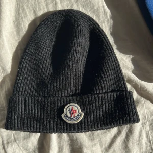 ÄKTA Svart ribbad mössa från Moncler - Snygg svart ribbad mössa från Moncler med broderad logotyp framtill. Perfekt för att hålla värmen under kyliga dagar. Tillverkad i ett mjukt och bekvämt material. Nypris 3000kr