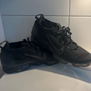 Svarta Nike Vapormax - Säljer ett par svarta Nike Vapormax sneakers med en unik sula och snörning. Skorna har en modern design och är perfekta för både träning och vardag. De är tillverkade i syntetmaterial för en lätt och bekväm känsla.                                         Storlek 42.      Snygga svarta skor👍.  Nypris ca2500kr