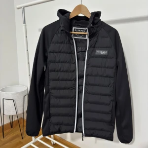 Svart cardigans från McKenzie - Snygg svart pufferjacka från McKenzie med dragkedja och huva. Jackan har en quiltad design och är perfekt för kyligare dagar. Den har en logotyp på bröstet och långa ärmar.