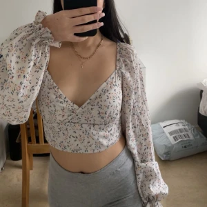 Blommig omlottopp med långa ärmar - Säljer en söt blommig omlottopp med långa puffärmar och v-ringning. Toppen är croppad och har ett romantiskt mönster i olika färger på en vit bakgrund. Perfekt för en somrig look!