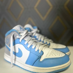 jordan 1 mid university blue - Snygga Nike Air Jordans i blått och vitt med grå detaljer. Skorna har klassisk snörning och perforerad tå för extra ventilation. Perfekta för dig som vill ha en stilren och sportig look. Kommer med original låda!