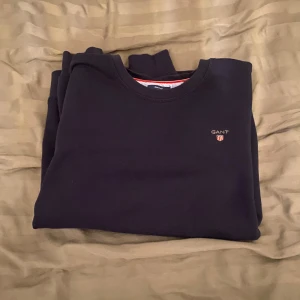 Blå tröja från GANT - Säljer en stilren blå tröja från GANT med rund halsringning och diskret logotyp på bröstet. Tröjan har långa ärmar och är perfekt för en avslappnad look.