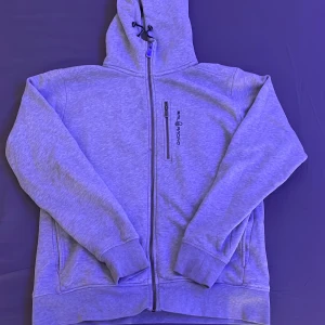 Grå hoodie från Sail Racing - Säljer en stilren grå hoodie från Sail Racing med dragkedja och justerbar huva. Tröjan har en praktisk bröstficka med dragkedja och ribbade muddar vid ärmslut och nederkant. Perfekt för en avslappnad stil.