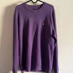 Lila tröja från Lyle & Scott - Säljer en snygg lila tröja från Lyle & Scott med rund hals och ett litet broderat emblem på bröstet. Tröjan har långa ärmar och ribbade muddar vid ärmslut och nederkant. Perfektk för en stilren look!