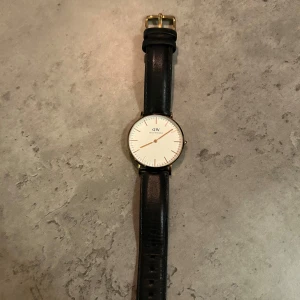 Klassisk klocka från Daniel Wellington - Säljer en stilren klocka från Daniel Wellington med svart läderarmband och vit urtavla. Klockan har en elegant guldfärgad boett och minimalistisk design. Perfekt för den som gillar tidlös stil.