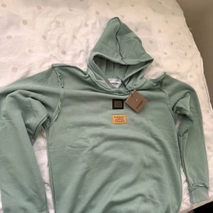 Grön hoodie från Burberry - Säljer en stilren grön hoodie från Burberry med märkets logga på bröstet. Hoodien har en klassisk design med långa ärmar och en bekväm huva. Perfekt för en avslappnad stil.