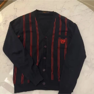 Svart kofta med röda ränder från Morris  - Snygg svart kofta från Morris med röda ränder och broderad logga på bröstet. Koftan har knappar framtill och långa ärmar, perfekt för en stilren look. Passar både till vardags och mer uppklädda tillfällen.