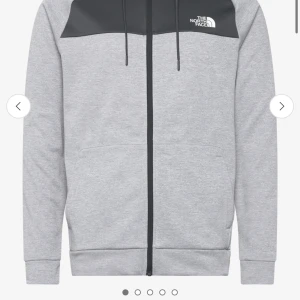 Grå hoodie från The North Face - Säljer en snygg grå hoodie från The North Face med svart panel upptill och dragkedja. Perfekt för dessa vår dagar med sin justerbara huva och bekväma passform. Loggan är tryckt på bröstet. Passar både för vardag och träning.
