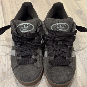 Bruna Adidas Campus sneakers - Snygga bruna Adidas Campus sneakers i mocka med klassiska vita ränder på sidan. Skorna har en robust brun sula och svarta snören. Perfekta för en stilren och bekväm look. Dem är helt oanvända. 