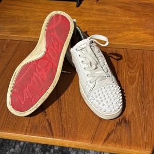 Vita Christian Louboutin  - Snygga vita sneakers med nitar på tån och klassisk röd sula. Skorna har snörning och är tillverkade i skinn, vilket ger en lyxig känsla. Perfekta för att ge din outfit en edgy touch. Väl andvända men fortfarande bra skick.