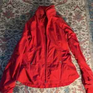 Röd vindjacka från Lululemon - Säljer en snygg röd vindjacka från Lululemon. Jackan har dragkedja framtill och praktiska fickor med dragkedjor. Perfekt för blåsiga dagar och ger en sportig look.