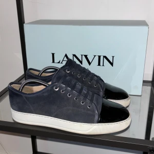  Lanvins - Mörkblå Lanvin med en stilren design. Skorna har en kombination av mocka och glansigt material på tån. De har snörning och en vit sula som ger en fräsch kontrast. Perfekta för en trendig look. Uk 8= 42/43