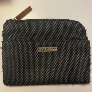 Svart plånbok från Giorgio Armani - Snygg svart plånbok från Giorgio Armani med en texturerad yta. Den har en dragkedja upptill och en metallplatta med logotypen på framsidan. Perfekt för att hålla dina kort och kontanter organiserade.