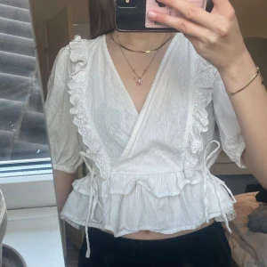 Vit omlottblus med spetsdetaljer - Charmig vit omlottblus med spetsdetaljer och volanger. Blusen har trekvartsärmar och knytband i midjan för en feminin touch. 