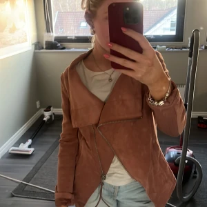 Rosa mockajacka med dragkedja - Snygg rosa mockajacka med asymmetrisk dragkedja och långärmad design. Perfekt för en stilren look med en touch av edge. Jackan har en unik omlottstil framtill.