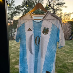 Argentina fotbollströja med nummer 10 - Säljer en costume made  Argentina fotbollströja  med nummer 10 och Messi på ryggen. Tröjan har de klassiska blå och vita ränderna och är kortärmad. Den har även ett emblem på bröstet och en hyllning till en legendarisk spelare Maradona vid högra sidan av tröjan (framifrån) hör av er vid minsta fundering eller fråga! 😁