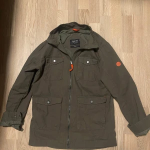 Olivgrön jacka från Indicode - Jag säljer den för ett billigare pris, oanvänd och helt ny.  Säljer en olivgrön jacka från Indicode med dragkedja och knappar. Jackan har fyra fickor framtill och en liten orange detalj på ärmen. Perfekt för höst och vår. 