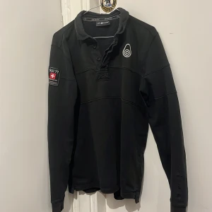 Svart pikétröja från Sail Racing - Snygg svart pikétröja från Sail Racing med coola detaljer som en broderad logga på bröstet och ett märke på ärmen med texten 'SUI 77 Extreme Sailing'. Perfekt för dig som gillar segling och stilren design. 