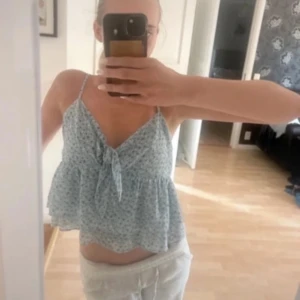 Blommig blå linnetopp - Säljer en söt blommig blå linnetopp med smala axelband och knytning framtill. Toppen har en luftig och lätt passform, perfekt för varma dagar.