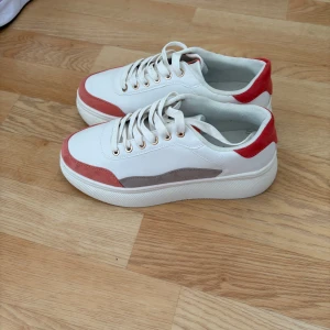 Vita sneakers med röda detaljer stl 39 - Snygga vita sneakers med röda och grå detaljer. Skorna har snörning och en bekväm sula som passar perfekt för vardagsbruk. De har en modern design med en kontrasterande färgkombination som ger en stilren look.