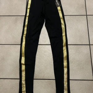 Svarta byxor/tajts  med guldglitter från Miss Mello - Snygga svarta byxor från Miss Mello med glittriga guldremsor längs sidorna. Perfekta för att sticka ut med en touch av glamour. Byxorna har en tight passform och är tillverkade i ett bekvämt syntetmaterial.