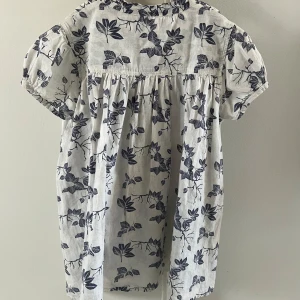 Vit och blå blommig blus från Bonton | storlek 140 - Charmig vit blus med blått blommönster från Bonton. Blusen har korta ärmar och knappar baktill. Perfekt för en somrig look!