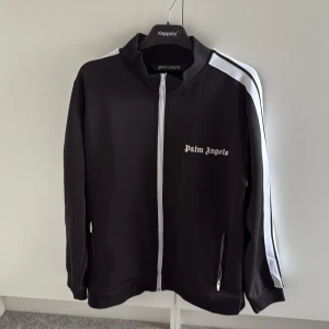 Svart jacka från Palm Angels - Snygg svart kodta från Palm Angels med vita detaljer och dragkedja framtill. Koftan har långa ärmar och två fickor med dragkedjor. Perfekt för en stilren look.