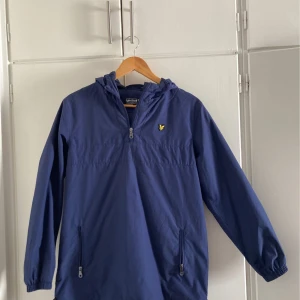 Blå vindjacka från Lyle & Scott - Snygg blå vindjacka från Lyle & Scott med dragkedja och huva. Jackan har en gul logotyp på bröstet och praktiska fickor med dragkedja. Perfekt för blåsiga dagar. Funkar för både tjejer och killar. Storlek 14/15y, passar för mig som vanligtvis bär storlek S. Kan diskutera pris