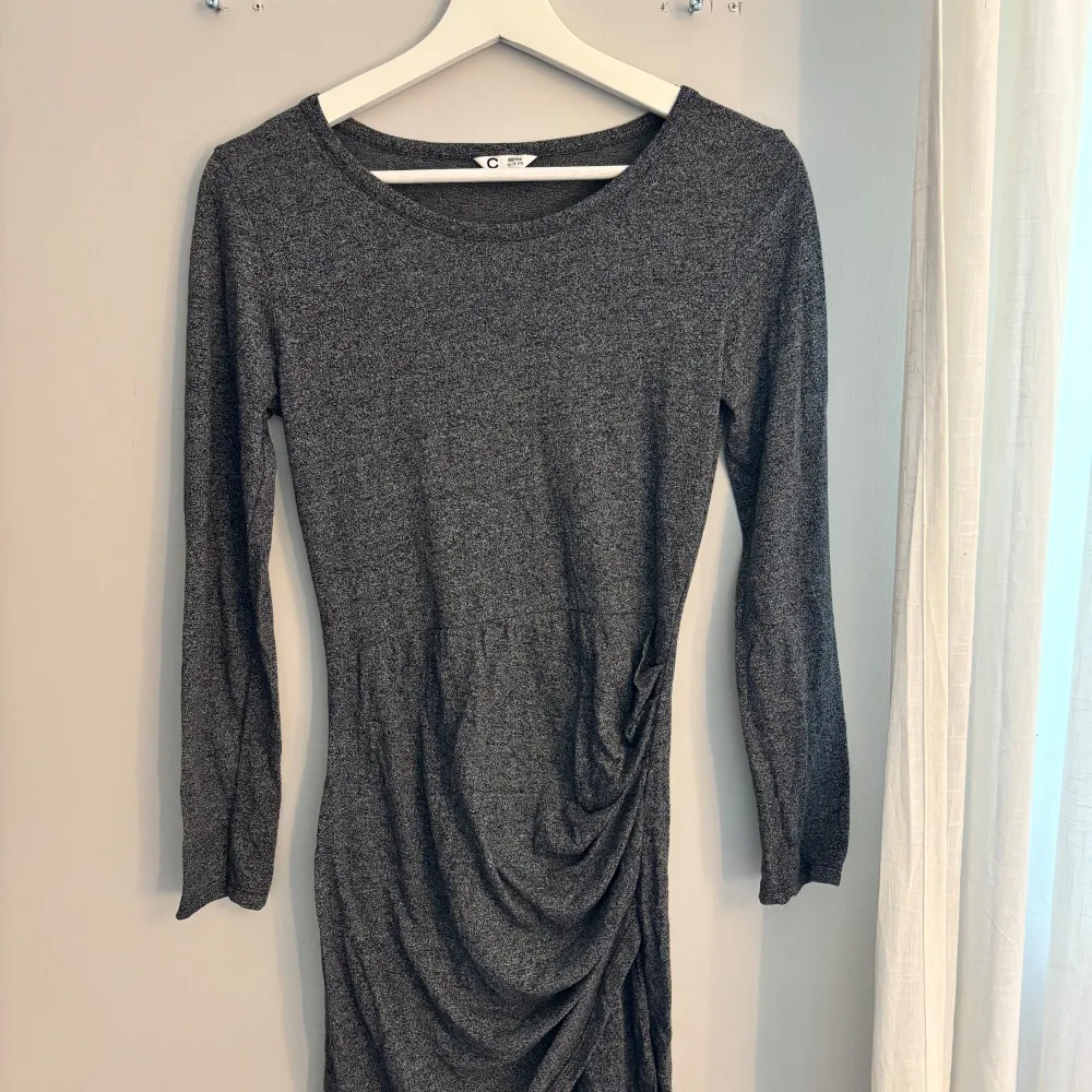 Snygg grå klänning med lång ärm och draperad detalj på sidan. Perfekt för en stilren look. Klänningen har en rund halsringning och är gjord i ett mjukt material.. Mekot.