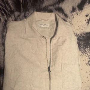 Beige skjorta med dragkedja från John Henric - Säljer en stilren beige skjorta från John Henric med dragkedja framtill. Skjortan har en klassisk krage och en bröstficka. Perfekt för en avslappnad men ändå uppklädd look. Storlek: M/L