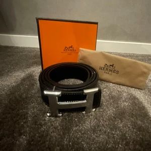 Svart bälte från H3rmes - Säljer ett stilrent svart bälte från H3rmes med en ikonisk H-formad spänne i silver. Skick 8/10 || storlek 115/46. Allt og ingår. Perfekt för att ge en lyxig touch till din outfit. Priset kan sänkas vid snabb affär.