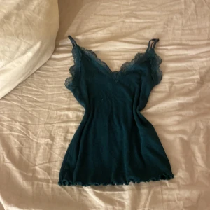 Mörkgrön ribbad linnetopp med spetsdetaljer  - Snygg mörkgrön ribbad linnetopp med spetsdetaljer vid halsringningen. Perfekt för en stilren och bekväm look. Justerbara axelband för bästa passform.