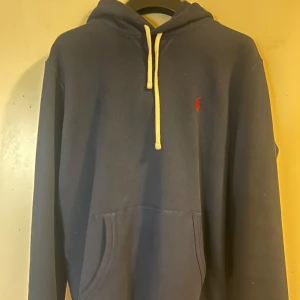 Mörkblå hoodie med broderad logga - Säljer en stilren mörkblå hoodie med en liten röd broderad logga på bröstet. Den har en klassisk känguruficka och vita snören i huvan. Perfekt för en avslappnad look. 10/10 skick, knappt använd! 
