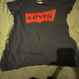 Svart t-shirt från Levi's - Säljer en svart t-shirt från Levi's med det klassiska röda logotyptrycket på framsidan. T-shirten har korta ärmar och en rund halsringning. Perfekt för en avslappnad stil.