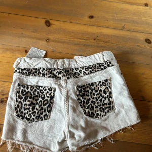 Vita shorts med leoparddetaljer - Snygga vita shorts med leopardmönstrade detaljer på fickorna och insidan av midjan. De har en fransig kant och knappar framtill. Perfekta för en cool och avslappnad stil.