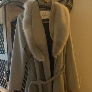 Grå kappa med pälskrage från Collection - Elegant grå kappa med en lyxig pälskrage och knytbälte i midjan. Perfekt för kyliga dagar när du vill känna dig både varm och stilren. Kappan har långa ärmar och knappar vid ärmsluten för extra detalj.